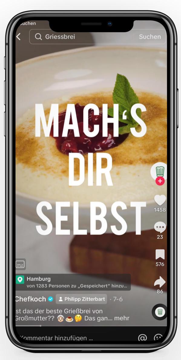 Das Bild zeigt ein Smartphone, auf dem ein Video abgespielt wird. Im Vordergrund ist eine Nahaufnahme eines Tellers mit Grießbrei zu sehen. Auf dem Grießbrei befinden sich eine rote Fruchtsauce und ein grünes Minzblatt als Dekoration. Der Bildschirm zeigt den Text 'MACH'S DIR SELBST', was dazu anregen soll, das Gericht selbst zuzubereiten. Die Benutzeroberfläche des Smartphones zeigt typische soziale Medien-Interaktionsmöglichkeiten wie Likes und Kommentare.
#!#
Grießbrei,Rezept,Selbstgemacht,Kochvideo,Fruchtsauce,Minze,Social Media,Smartphone,Kochen,Anleitung