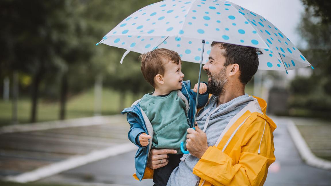 Ein Vater und sein kleiner Sohn stehen lächelnd unter einem blauen Regenschirm mit weißen Punkten. Der Vater trägt eine gelbe Regenjacke und hält den Jungen, der eine blaue Jacke trägt. Der Hintergrund zeigt eine regnerische Parklandschaft mit Bäumen und einem leeren Parkplatz.
#!#
Vater,Sohn,Regen,Regenschirm,Familie,Gelbe Jacke,Blauer Schirm,Lächeln,Natur,Elternschaft