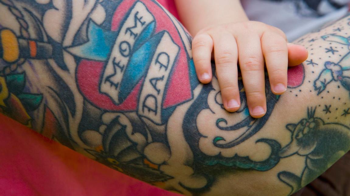 Das Bild zeigt eine Nahaufnahme eines tätowierten Arms, auf dem ein Herz mit den Worten 'Mom' und 'Dad' sichtbar ist. Eine kleine Kinderhand berührt sanft den Arm, was eine Verbindung zwischen Familienliebe und Generationen symbolisiert. Der Arm ist mit farbenfrohen Tattoos verziert, die verschiedene Muster und Designs umfassen.
#!#
Tätowierung,Familie,Herz,Kind,Liebe,Berührung,Muster,Generationen,Farben,Symbolik