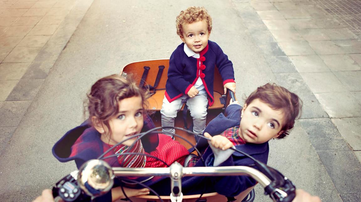 Drei kleine Kinder sitzen in einer großen Kiste vorne auf einem Fahrrad, das auf einer gepflasterten Straße fährt. Die Kinder tragen warme Kleidung in Blau und Rot und schauen neugierig in die Kamera. Die Kiste ist aus Holz und das Fahrrad wird von einer nicht sichtbaren Person gesteuert. Die Umgebung ist städtisch mit einer gepflasterten Straße im Hintergrund.
#!#
Kinder,Fahrrad,Stadt,Kiste,Spaß,Abenteuer,Familie,Transport,Straße,Neugier