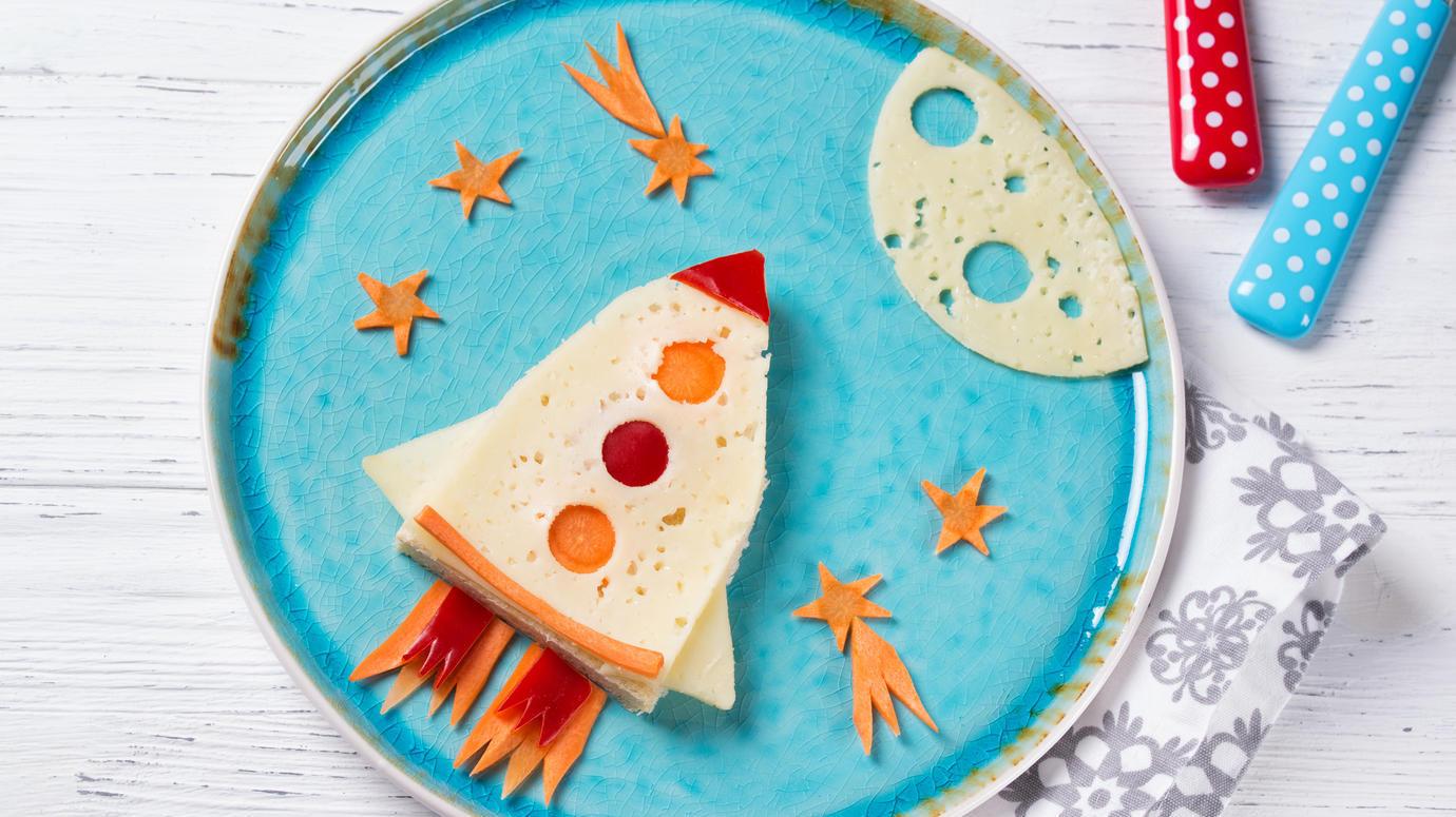 Das Bild zeigt einen kreativ gestalteten Teller mit einer aus Käse, Brot und Gemüse geformten Rakete. Die Rakete besteht aus einer Scheibe Käse mit runden Ausstanzungen, die von Karotten- und Paprikastückchen begleitet wird. Die Szenerie ist mit einem Mond aus Käse und kleinen Sternen aus Karotten auf einem blauen Teller dekoriert. Bunte Besteckteile und eine gemusterte Serviette ergänzen das Arrangement.
#!#
Kinder,Essen,Kreativität,Rakete,Weltraum,Käse,Gemüse,Brot,Dekoration,Snacks