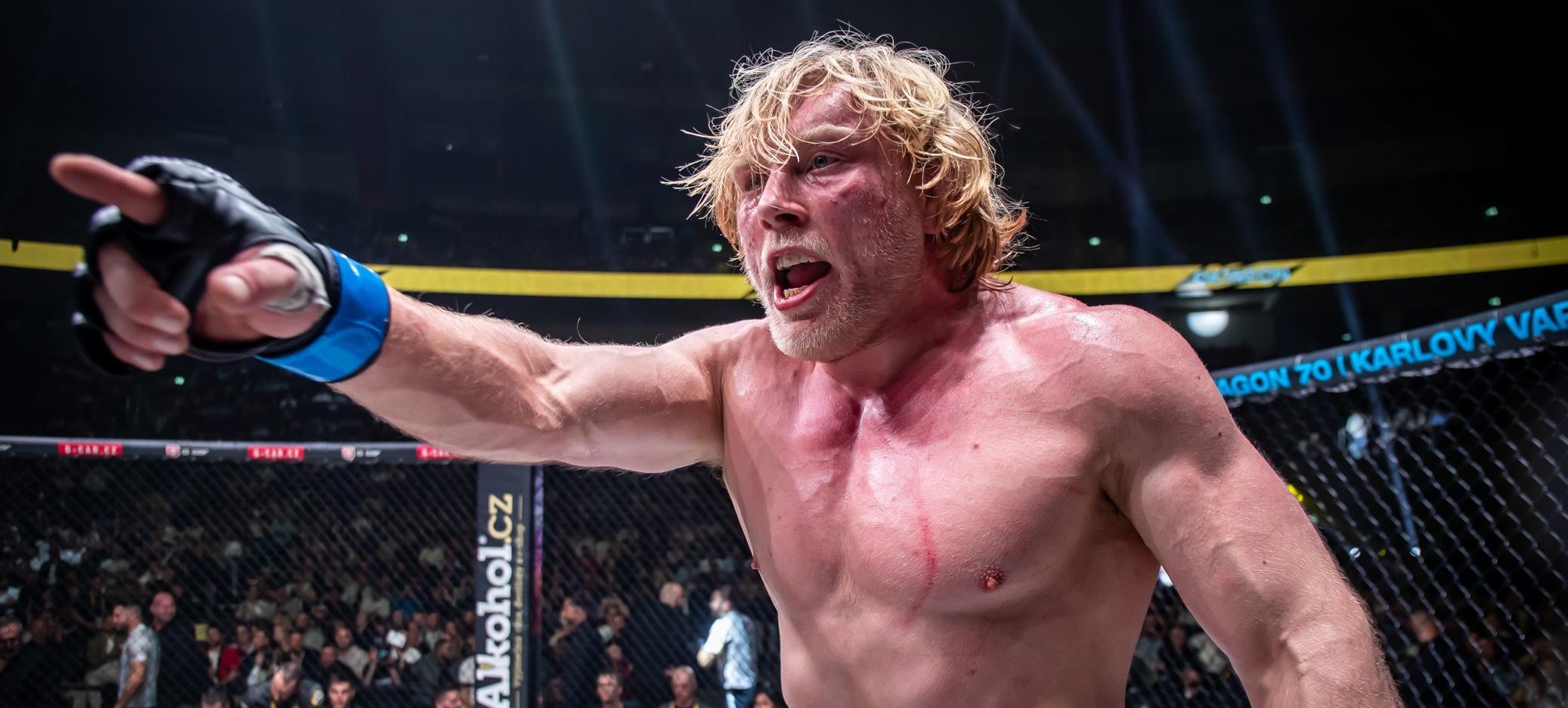 Ein muskulöser, blondhaariger Kämpfer steht im Mittelpunkt eines MMA-Kampfes in einem Oktagon. Er trägt schwarze Handschuhe und einen blauen Handgelenkschutz, während er entschlossen und schweißbedeckt in Richtung seines Gegners zeigt. Der Hintergrund zeigt ein aufmerksames Publikum, das den Kampf beobachtet, sowie einen Käfig, der den Ring umgibt. Lichter und Banner sind über der Kampfarena sichtbar.
#!#
MMA,Kämpfer,Oktagon,Kampf,Sport,Publikum,Intensität,Handschuhe,Käfig,Arena