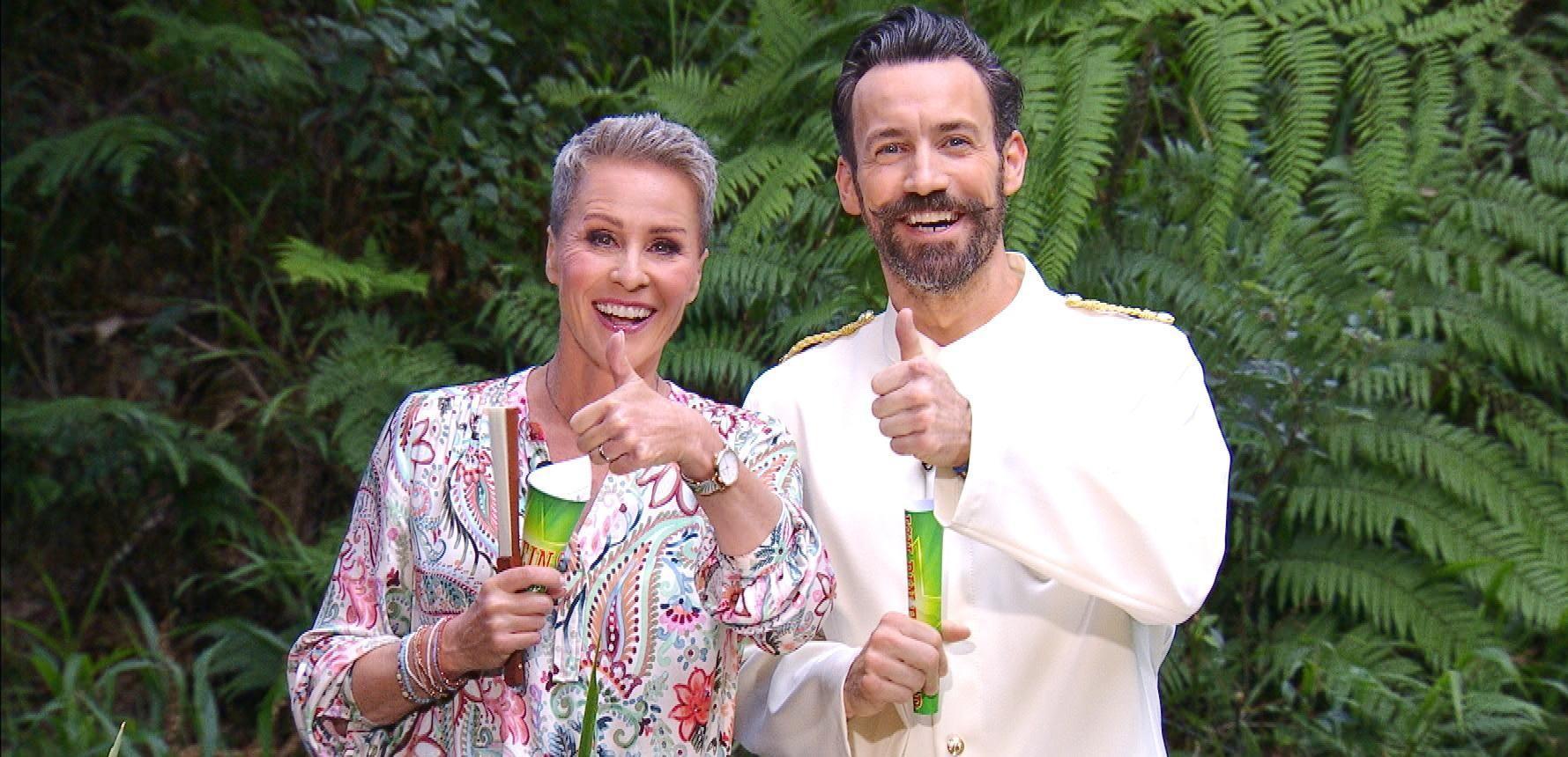 Zwei Personen stehen lächelnd in einem üppigen, grünen Dschungel. Die Frau trägt eine gemusterte Bluse und hält ein Mikrofon, während der Mann in einem weißen Anzug ebenfalls ein Mikrofon hält. Beide zeigen mit einem Daumen nach oben und wirken sehr fröhlich und entspannt. Im Hintergrund sind dichte Farne und andere tropische Pflanzen zu sehen.
#!#
Dschungel,Lächeln,Mikrofon,Daumen hoch,Tropisch,Grün,Bluse,Anzug,Natur,Fröhlichkeit