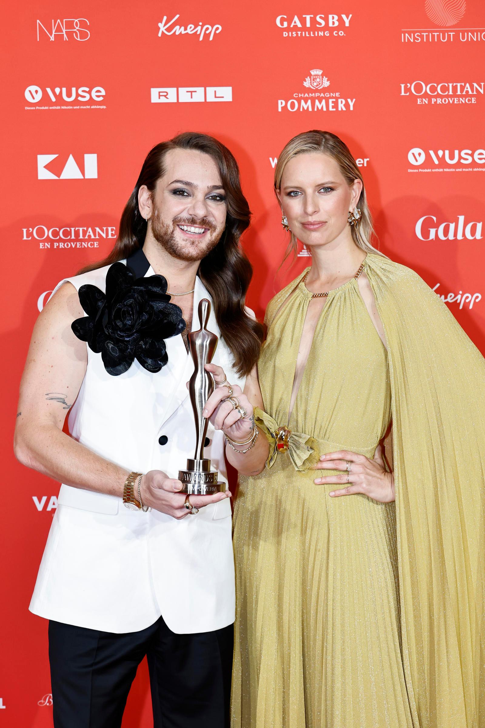 Das Bild zeigt zwei elegant gekleidete Personen auf einem roten Teppich bei einer Preisverleihung. Die Person links trägt ein weißes Sakko mit einer großen schwarzen Blume als Dekoration und hält eine Trophäe in der Hand. Die Person rechts trägt ein bodenlanges, gelbes Kleid mit einem Umhang. Beide Personen lächeln in die Kamera und stehen vor einer Sponsorenwand mit verschiedenen Logos.
#!#
Preisverleihung,roter Teppich,Glamour,Mode,Trophäe,Sponsorenwand,Eleganz,Sakko,Kleid,Lächeln