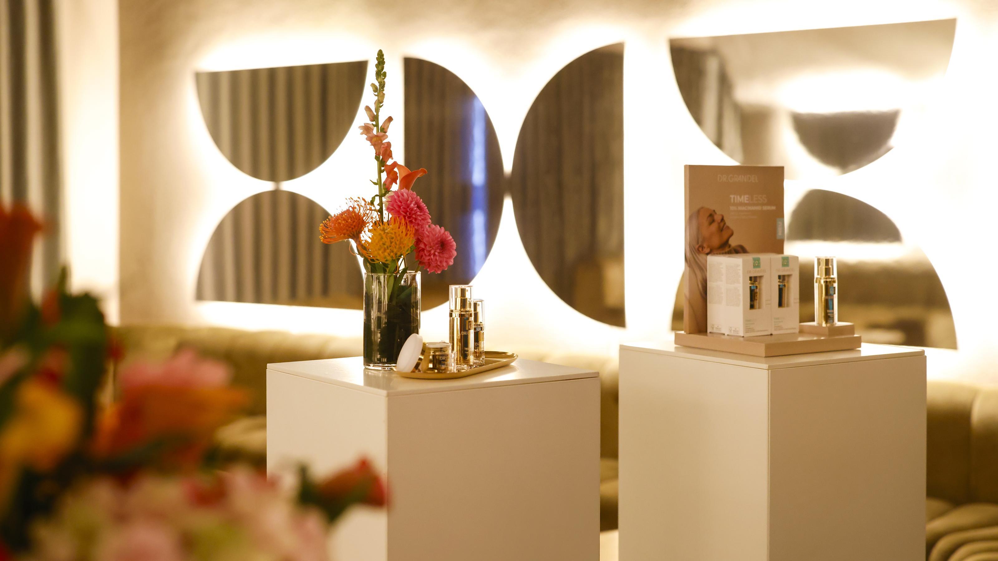 Das Bild zeigt einen stilvoll eingerichteten Raum mit zwei weißen Podesten im Vordergrund. Auf dem linken Podest steht eine Vase mit bunten Blumen und daneben luxuriöse Pflegeprodukte in goldenen Flaschen. Das rechte Podest ist mit einer Broschüre und weiteren Pflegeprodukten dekoriert. Im Hintergrund befinden sich große, dekorative Spiegel, die das Licht reflektieren und dem Raum eine elegante Atmosphäre verleihen.
#!#
Luxus,Pflegeprodukte,Blumen,Spiegel,Dekoration,Innenraum,Eleganz,Beleuchtung,Präsentation,Ästhetik