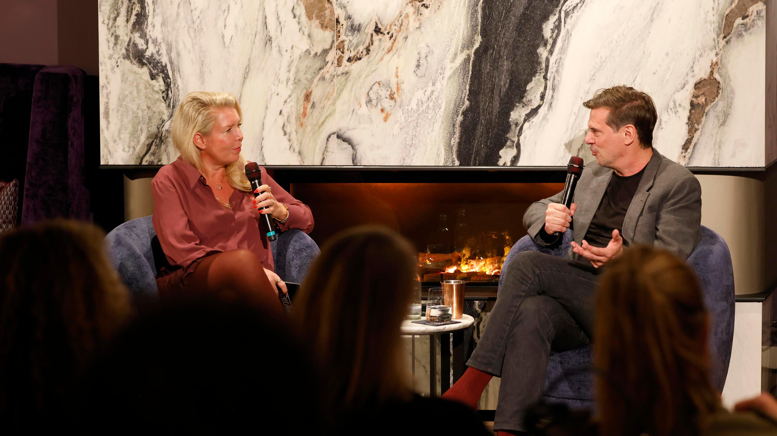 Das Bild zeigt zwei Personen, die in einem modernen, gemütlichen Raum auf blauen Sesseln sitzen und sich unterhalten. Beide halten Mikrofone in der Hand, was auf ein Interview oder eine Diskussion hinweist. Im Hintergrund ist ein stilvoller Kamin mit einer weißen, marmorartigen Verkleidung zu sehen, der eine warme Atmosphäre schafft. Im Vordergrund sind unscharfe Köpfe von Zuschauern zu erkennen, die dem Gespräch folgen.
#!#
Interview,Diskussion,Kamin,Mikrofon,Zuhörer,Gespräch,Event,Moderator,Publikum,Atmosphäre