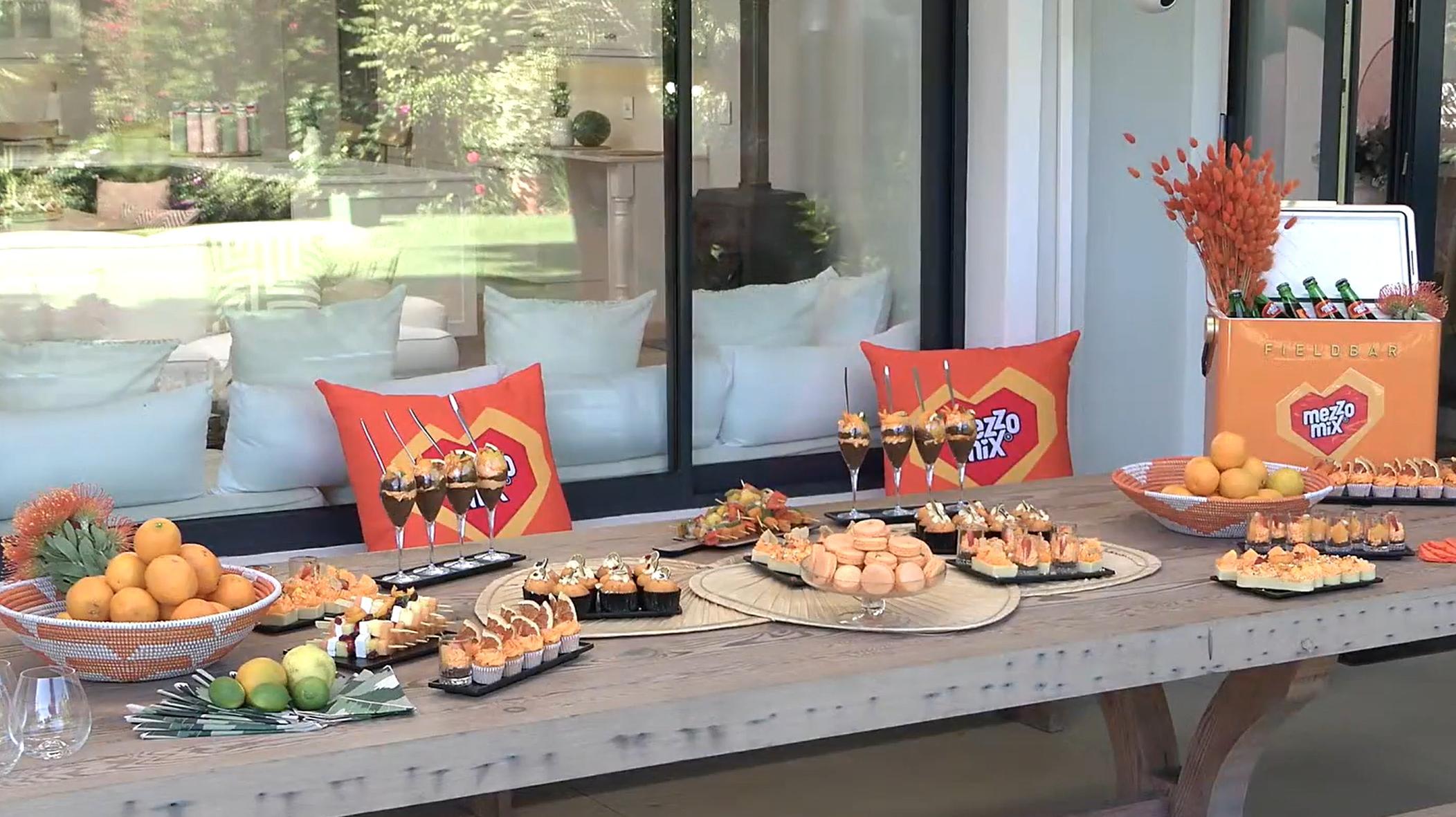 Das Bild zeigt einen liebevoll dekorierten Tisch, der im Freien für eine sommerliche Gartenparty vorbereitet ist. Auf dem Tisch befinden sich zahlreiche Teller und Platten mit verschiedenen Snacks, die alle in einem orangefarbenen und frischen Farbschema gehalten sind. Im Hintergrund sind zwei orangefarbene Kissen mit dem Mezzo Mix Logo zu sehen, die auf einer weißen Sitzgelegenheit platziert sind. Rechts steht eine Kühlbox mit Getränken, die ebenfalls das Mezzo Mix Logo trägt. Der Tisch ist mit Früchten wie Orangen und Zitronen dekoriert und vermittelt eine sommerlich-fröhliche Atmosphäre.
#!#
Gartenparty,Mezzo Mix,Snacks,Sommer,Dekoration,Orangen,Getränke,Kissen,Früchte,Kühlbox
