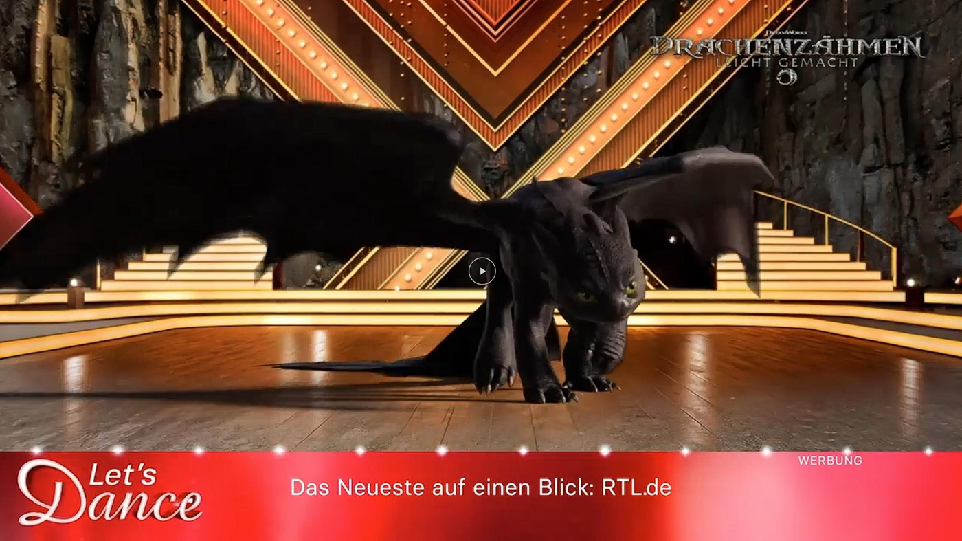 Ein animierter schwarzer Drache mit großen Flügeln steht auf einer hell erleuchteten Bühne mit goldenen und roten Lichtelementen im Hintergrund. Die Bühne hat eine elegante Treppe, die zu einem dunklen, steinernen Hintergrund führt, der an eine Höhle erinnert. Unten ist ein roter Banner mit dem Logo von 'Let's Dance' und der Website RTL.de zu sehen.
#!#
Drache,Animation,Let’s Dance,Bühne,RTL,Lichter,Hintergrund,Treppe,Höhle,Werbung